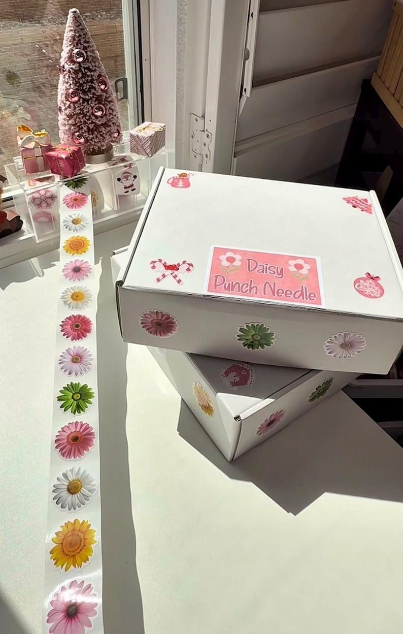 May include: Two white boxes adorned with daisy stickers and a label reading "Daisy Punch Needle." A strip of daisy stickers and a small pink Christmas tree are visible. The boxes are stacked on a white surface.