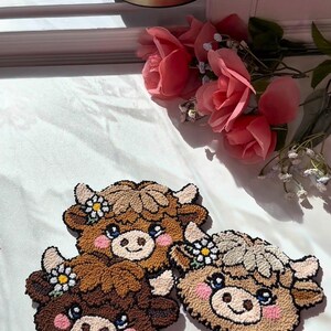 Op de afbeelding: Drie handgemaakte decoratieve items in de vorm van een koe. Elk heeft een bruin, beige en cr&egrave;mekleurig kleurenschema, met een lachend gezicht, roze wangen en een bloemenkrans. De items zijn gemaakt met een getextureerde, geluste garentechniek.