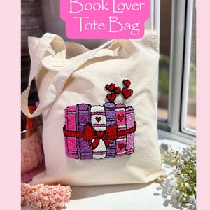May include: A cream tote bag with the text "Book Lover Tote Bag" in a pink rectangle. The bag features an embroidered design of books in pink and purple, tied with a red bow and topped with red hearts.