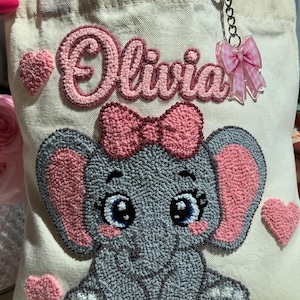 May include: A canvas tote bag with pink handles and accents. The bag features a gray elephant design with pink ears and a bow, along with the name "Olivia" in pink. Pink heart accents and a bow-shaped charm complete the design.