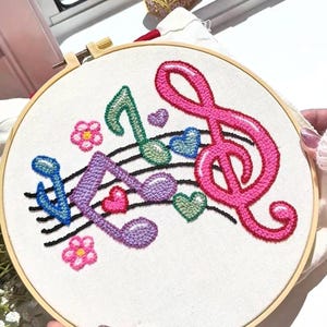 May include: Embroidered musical notes and treble clef in pink, green, blue, and purple on a white fabric stretched in a wooden hoop. The design includes hearts and flowers.