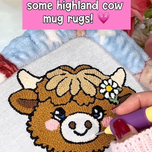 May include: A Highland cow mug rug is being punch needled. The rug features a brown cow face with a flower and the text "Let's punch needle some highland cow mug rugs!" on a pink background.