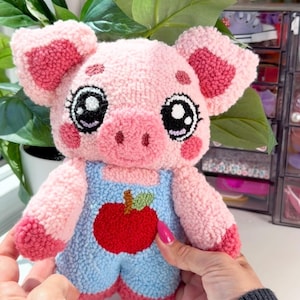 May include: A plush pig toy crafted with punch needle texture, featuring a light pink body, dark pink ear accents, and large black and white eyes. The pig wears light blue overalls with a prominent red apple graphic on the front chest area.