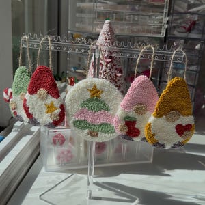 May include: A display of handmade Christmas ornaments. The ornaments include gnome and Christmas tree designs, crafted with textured, colorful yarn. Each ornament is suspended by twine and displayed on a clear acrylic stand.