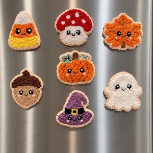 Könnte beinhalten: Ein Set aus sieben Punch-Needle-Magneten mit niedlichen, lächelnden Gesichtern. Die Designs umfassen einen Zuckermais, einen rot-weißen Pilz, ein orangefarbenes Ahornblatt, eine braune Eichel, einen orangefarbenen Kürbis, ein weißes Gespenst und einen lila Hexenhut.