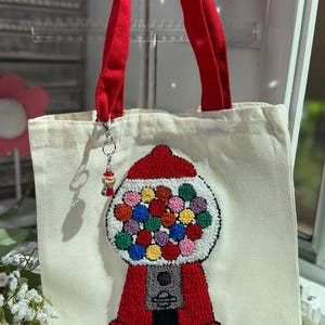 Può includere: Borsa tote in tela color crema con un design a distributore di palline rosse. Il distributore è rosso con la parte superiore bianca e pieno di palline colorate. La borsa ha manici rossi e un piccolo ciondolo a distributore.
