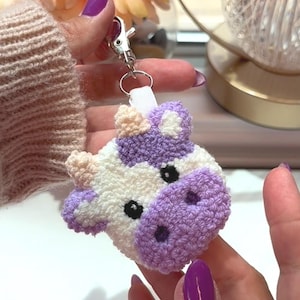 May include: A handmade, crocheted cow keychain in white, lavender, and light beige. The cow's face is detailed with black eyes and a lavender muzzle. The keychain has a silver-colored clasp and ring.