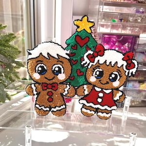 May include: Handmade Christmas ornaments featuring a gingerbread man and woman, and a Christmas tree. The gingerbread figures have white frosting hair and accents, with red bow ties and dresses. The tree is green with a yellow star and red heart accents.