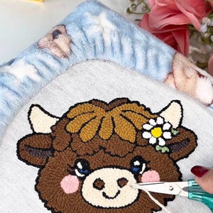 May include: A close-up of a brown and tan cow face design on a light grey fabric. The cow has blue eyes, pink cheeks, and a daisy flower crown. Scissors are cutting a thread from the design. A blue blanket with bear designs is in the background.