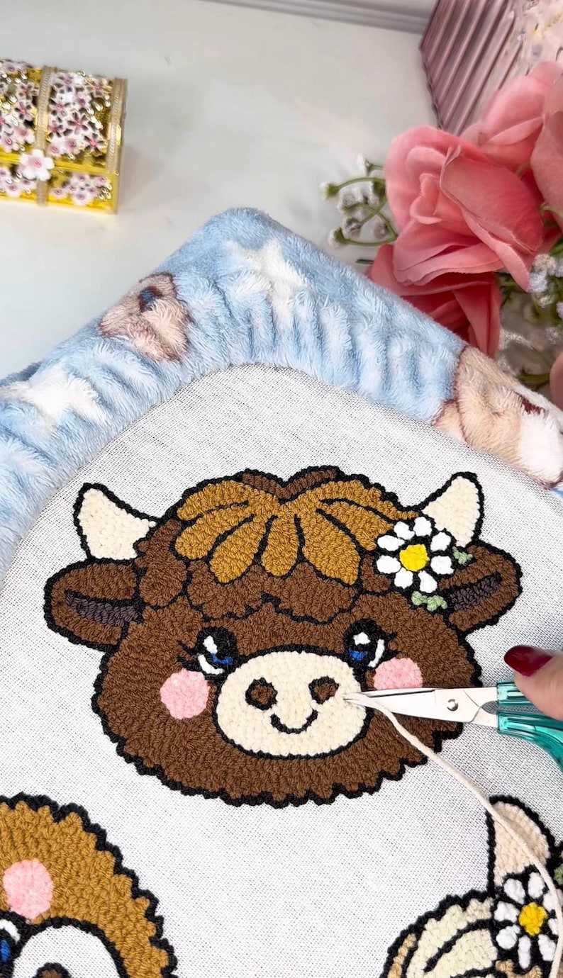 May include: A close-up of a brown and tan cow face design on a light grey fabric. The cow has blue eyes, pink cheeks, and a daisy flower crown. Scissors are cutting a thread from the design. A blue blanket with bear designs is in the background.