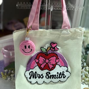 Può includere: Borsa tote in tela color crema con manici rosa e un portachiavi a pompon rosa. La borsa presenta un colorato design ricamato di una mela, un fiocco e un arcobaleno, con il nome "Mrs Smith" in grafia nera.