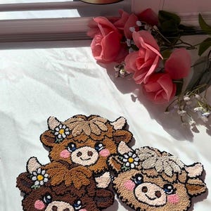 May include: Three handmade cow-shaped decorative items. Each has a brown, tan, and cream colour scheme with a smiling face and a flower crown. The items are made of a textured material, possibly yarn or fabric, and are placed on a white surface.