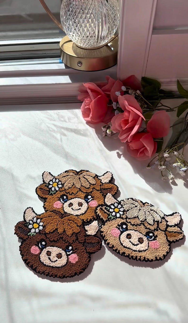 May include: Three handmade cow-shaped decorative items. Each has a brown, tan, and cream colour scheme with a smiling face and a flower crown. The items are made of a textured material, possibly yarn or fabric, and are placed on a white surface.
