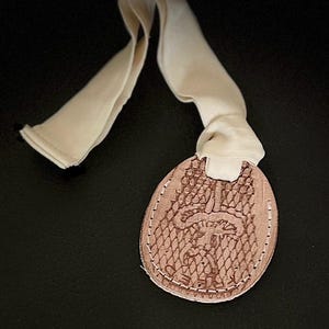 Puede incluir: Una etiqueta de cuero marrón claro con un borde cosido y un diseño de hongo. La etiqueta está unida a un lazo de tela blanco.