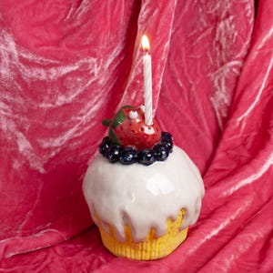May include: A white ceramic cupcake topped with a strawberry and blueberries. A lit white candle sits in the centre of the cupcake. The cupcake is resting on a pink velvet surface.