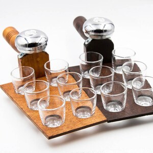 May include: A wooden serving tray with two attached metal and wood handles, each holding a set of clear shot glasses. The tray is divided into two sections, one light brown and one dark brown, each holding six shot glasses.