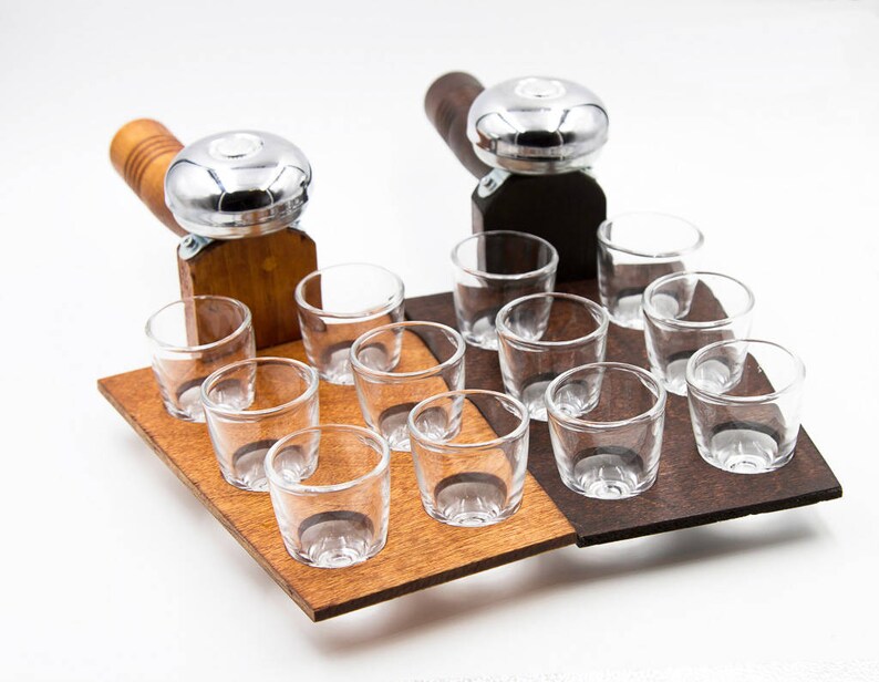 May include: A wooden serving tray with two attached metal and wood handles, each holding a set of clear shot glasses. The tray is divided into two sections, one light brown and one dark brown, each holding six shot glasses.
