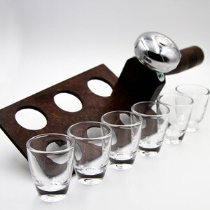May include: A wooden shot glass holder with six clear shot glasses. The holder is dark brown with three circular cutouts. A silver-colored metal bottle opener is attached to the holder.