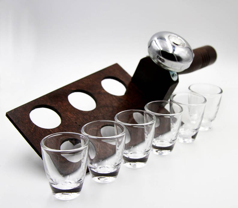 May include: A wooden shot glass holder with six clear shot glasses. The holder is dark brown with three circular cutouts. A silver-colored metal bottle opener is attached to the holder.