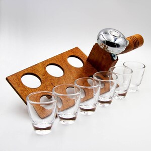 May include: A wooden shot glass holder with six clear shot glasses. The holder has a built-in bell and a wooden handle. The shot glass holder is made of dark brown wood and has a rectangular shape with circular cutouts.