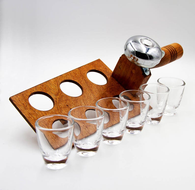 May include: A wooden shot glass holder with six clear shot glasses. The holder has a built-in bell and a wooden handle. The shot glass holder is made of dark brown wood and has a rectangular shape with circular cutouts.