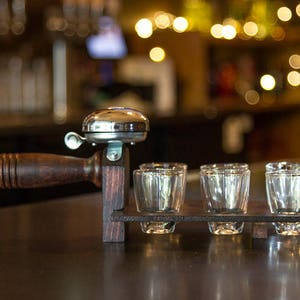 May include: A wooden shot glass holder with a silver bell and four clear shot glasses. The holder has a dark brown handle and a rectangular base. The background is blurred with warm lighting.