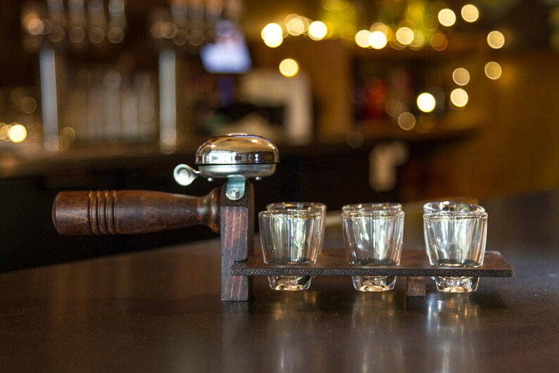 May include: A wooden shot glass holder with a silver bell and four clear shot glasses. The holder has a dark brown handle and a rectangular base. The background is blurred with warm lighting.