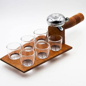 Könnte beinhalten: Ein Holz-Shotglashalter mit sechs klaren Shotgläsern. Der Halter hat eine eingebaute Glocke mit einem Holzgriff. Das Holz hat einen warmen, braunen Ton und die Gläser sind leer. Die Glocke ist silberfarben.
