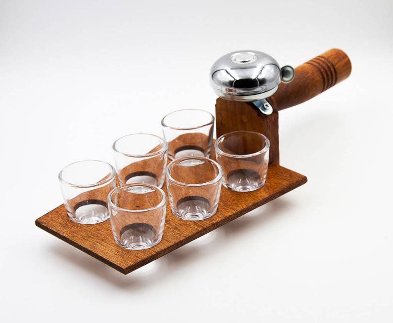 May include: A wooden shot glass holder with six clear shot glasses. The holder has a built-in bell with a wooden handle. The wood has a warm, brown tone, and the glasses are empty. The bell is silver.