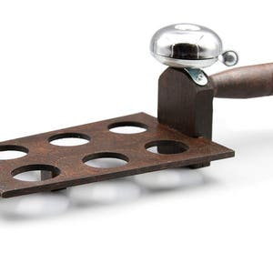 May include: A wooden shot glass holder with six circular openings. It has a dark brown finish and is paired with a silver bell and a wooden handle. The bell is mounted on a wooden support.