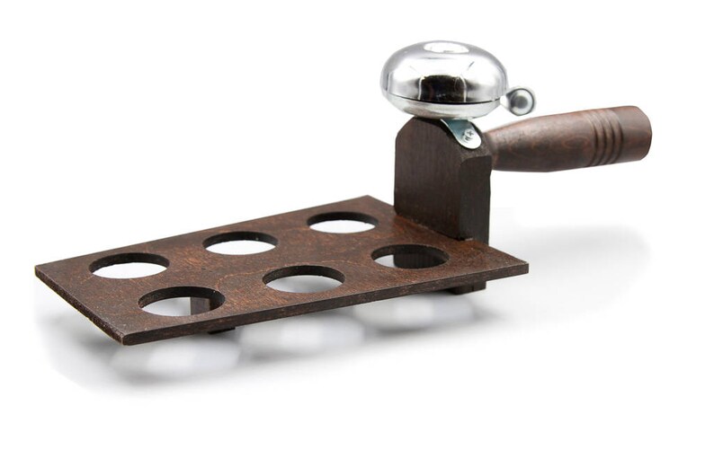 May include: A wooden shot glass holder with six circular openings. It has a dark brown finish and is paired with a silver bell and a wooden handle. The bell is mounted on a wooden support.