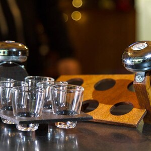 May include: A set of shot glasses on a dark gray slate tray, alongside a wooden holder with four circular cutouts. A shiny chrome bell is attached to a wooden handle, adding a decorative touch to the barware.