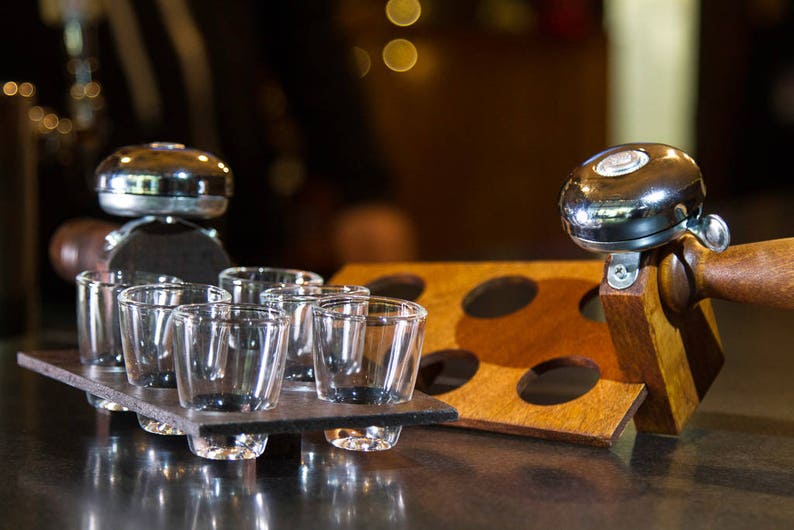 May include: A set of shot glasses on a dark gray slate tray, alongside a wooden holder with four circular cutouts. A shiny chrome bell is attached to a wooden handle, adding a decorative touch to the barware.