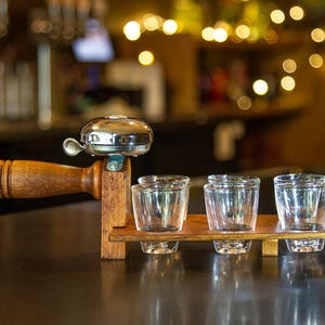May include: A wooden shot glass holder with a silver bell and four clear shot glasses. The holder has a dark brown finish and is designed to be held by a wooden handle. The background is blurred, suggesting a bar setting.