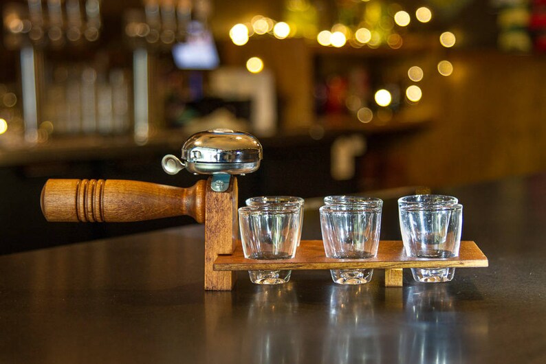 May include: A wooden shot glass holder with a silver bell and four clear shot glasses. The holder has a dark brown finish and is designed to be held by a wooden handle. The background is blurred, suggesting a bar setting.