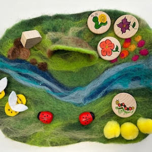 May include: A felt play mat with a green field, a blue river, and a brown hill. There are wooden cutouts of a snail, a flower, a ladybug, a bee, and a caterpillar. There are also yellow pom-poms and a small wooden house.