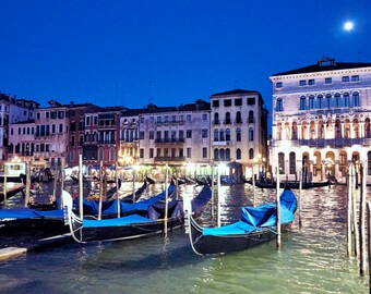 Puede incluir: Una vista nocturna de un canal en Venecia, Italia, con varias góndolas atracadas a lo largo del borde. Las góndolas son de color azul oscuro con ribetes negros y tienen toldos azules. Los edificios a lo largo del canal están iluminados y se reflejan en el agua. Se puede ver una luna llena en el cielo.