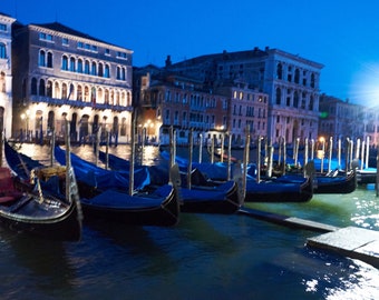 Puede incluir: Una fila de góndolas están atracadas en un muelle en Venecia, Italia. Las góndolas son negras con cubiertas de tela azul y están alineadas en una fila. Las góndolas están frente a una fila de edificios con luces encendidas. El cielo es azul oscuro y el agua está tranquila.