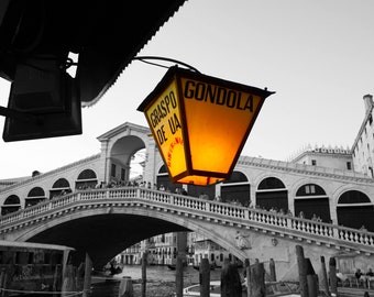 Puede incluir: Una linterna amarilla con las palabras "GONDOLA" y "GRASPO DE UA" en ella, colgada de un soporte de metal negro. La linterna está encendida y brilla, proyectando una luz cálida. La linterna está delante de una foto en blanco y negro de un puente en Venecia, Italia.