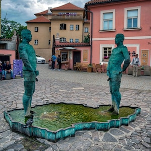 Puede incluir: Dos estatuas verdes de hombres de pie en una plaza adoquinada frente a un edificio rosa. Las estatuas forman parte de una fuente con una cuenca de agua verde en forma de mapa.