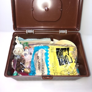 Puede incluir: Una caja de almacenamiento de plástico marrón con tapa está abierta y llena de diversos artículos de costura, incluyendo una bolsa de parches de bolsillo termoadhesivos, una bobina de hilo, botones y un ribete de encaje blanco. La bolsa de parches de bolsillo termoadhesivos tiene el texto "IRON-ON POCKET PATCHES" y "ANTI AND JACKETS" impreso en ella.