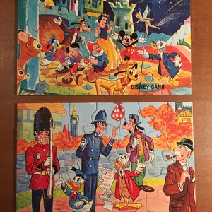 May include: Two vintage jigsaw puzzles featuring Disney characters. The first puzzle shows Mickey Mouse, Donald Duck, Goofy, Snow White, and other Disney characters in front of a castle. The second puzzle shows Mickey Mouse, Donald Duck, Goofy, and other Disney characters in front of a building with a sign that says "A Guide".