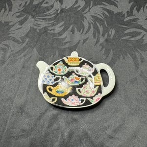 May include: A white ceramic teapot-shaped tea bag holder with a black background. The holder is decorated with various colorful teapots. The holder has a handle and a small spout. The holder is approximately 5 inches long and 4 inches wide.