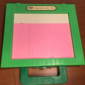 Puede incluir: Kit de cuentos de plástico verde con asa y una etiqueta blanca que dice "Storybook Kit". El kit tiene tres paneles rosas con diseños en relieve.