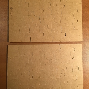 May include: Two brown cardboard puzzle pieces with a jigsaw pattern. The pieces are blank and ready to be used for a custom puzzle.