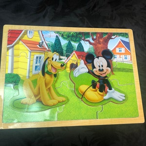 Puede incluir: Un rompecabezas de madera con Mickey Mouse y Pluto. Mickey lleva pantalones cortos rojos y zapatos amarillos. Pluto es un perro amarillo con un collar verde. El rompecabezas está ambientado en un fondo de una casa y un césped verde.