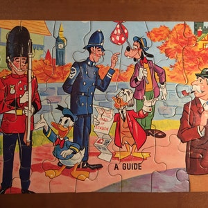 May include: A vintage jigsaw puzzle featuring cartoon characters like Donald Duck, Goofy, and a British guard. The puzzle depicts a scene in London with a red double-decker bus, a map of London, and a guidebook. The puzzle is made of cardboard and has a colorful design.