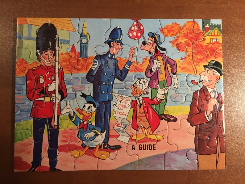May include: A vintage jigsaw puzzle featuring cartoon characters like Donald Duck, Goofy, and a British guard. The puzzle depicts a scene in London with a red double-decker bus, a map of London, and a guidebook. The puzzle is made of cardboard and has a colorful design.
