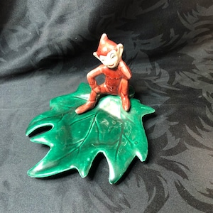 Könnte beinhalten: Eine Keramikfigur eines roten Kobolds, der auf einer grünen, blattartigen Schale sitzt. Der Kobold trägt eine spitze Mütze und hält sich die Hand ans Ohr.