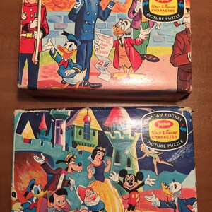 May include: Two vintage Walt Disney character picture puzzles in their original boxes. The top box features Donald Duck, a British policeman, and a man with a map of London. The bottom box features Mickey Mouse, Snow White, and other Disney characters.